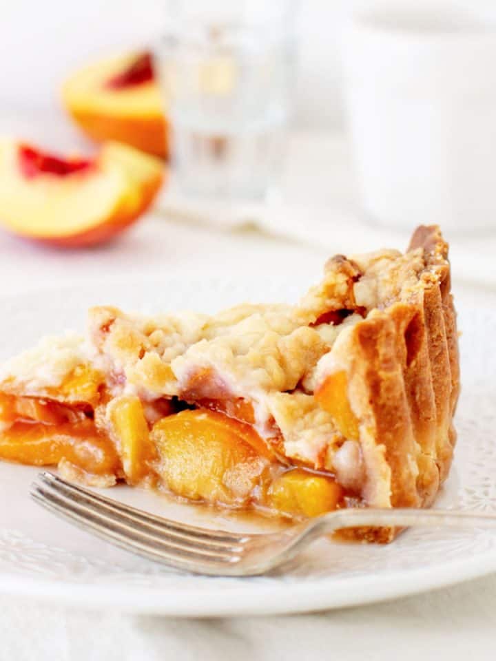 Peach crumb pie slice on a white plate with silver fork, a white background.