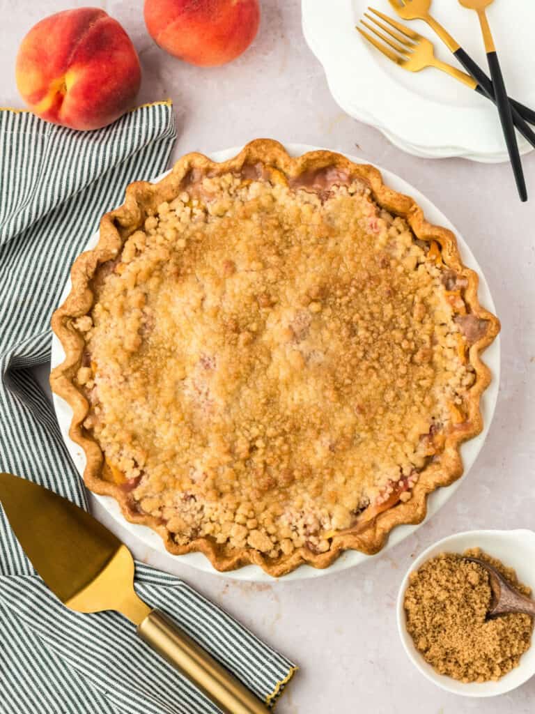 Baked crumb peach pie. Green towel, gold utensils, whitish surface. Peaches.