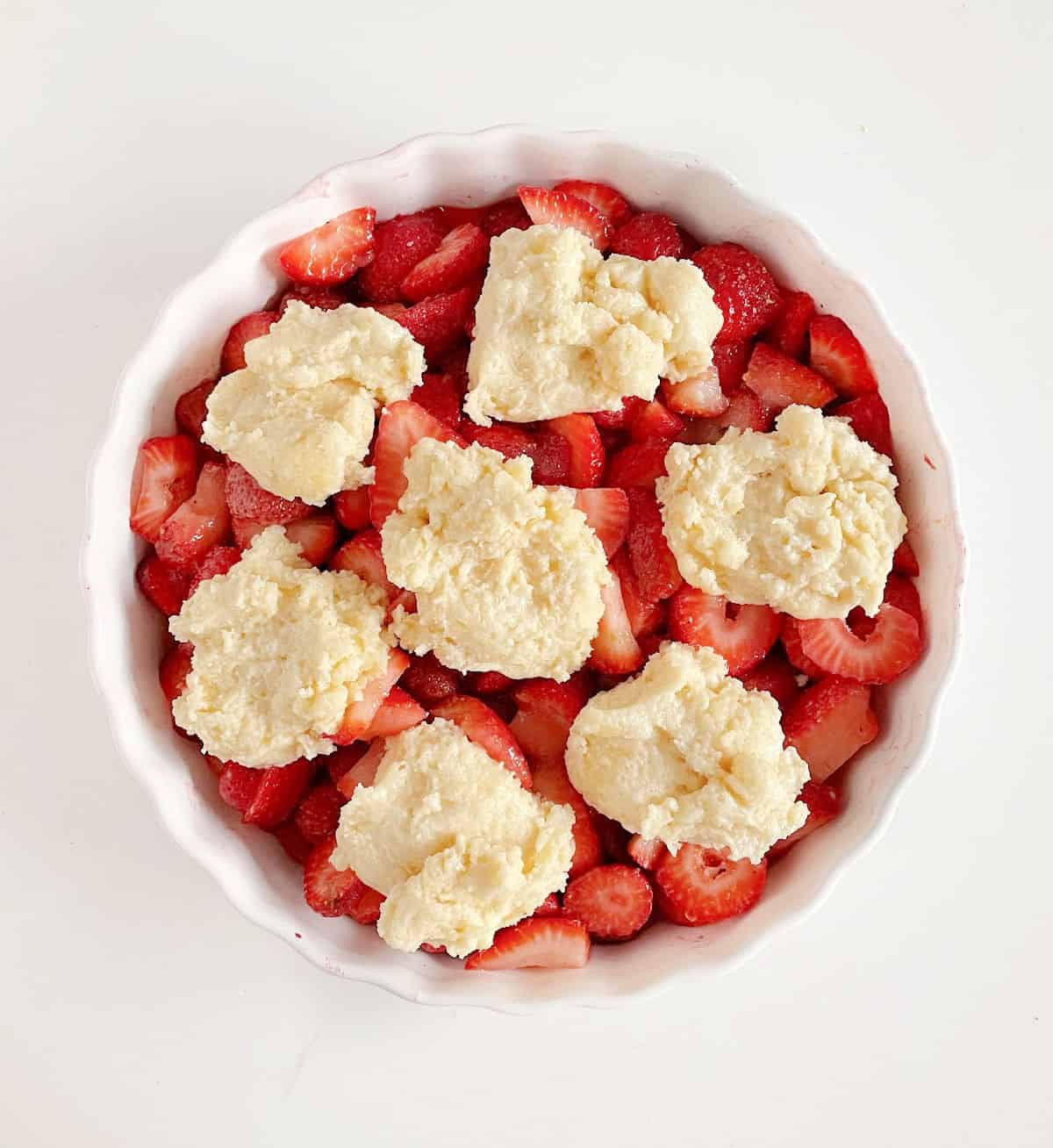 Overview of round white ceramic dish with unbaked straberry cobbler on white surface.