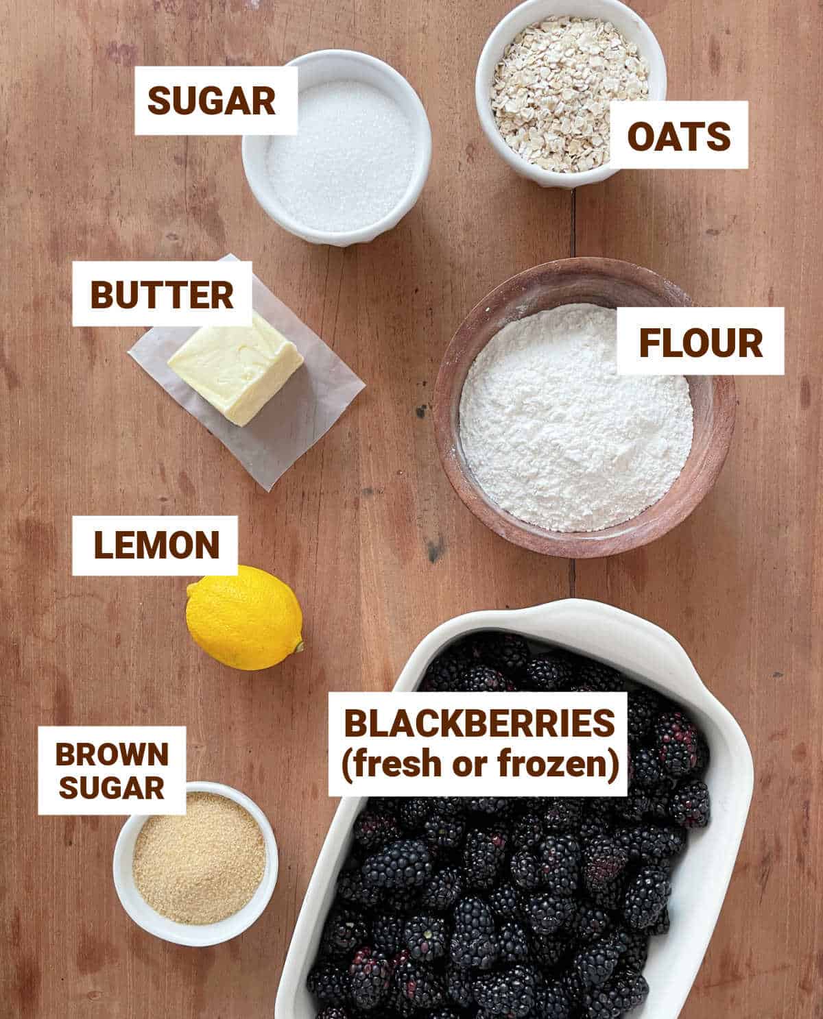 Ingredients for blackberry crisp in bowls on a wooden table including lemon, sugar, butter, flour, oats.