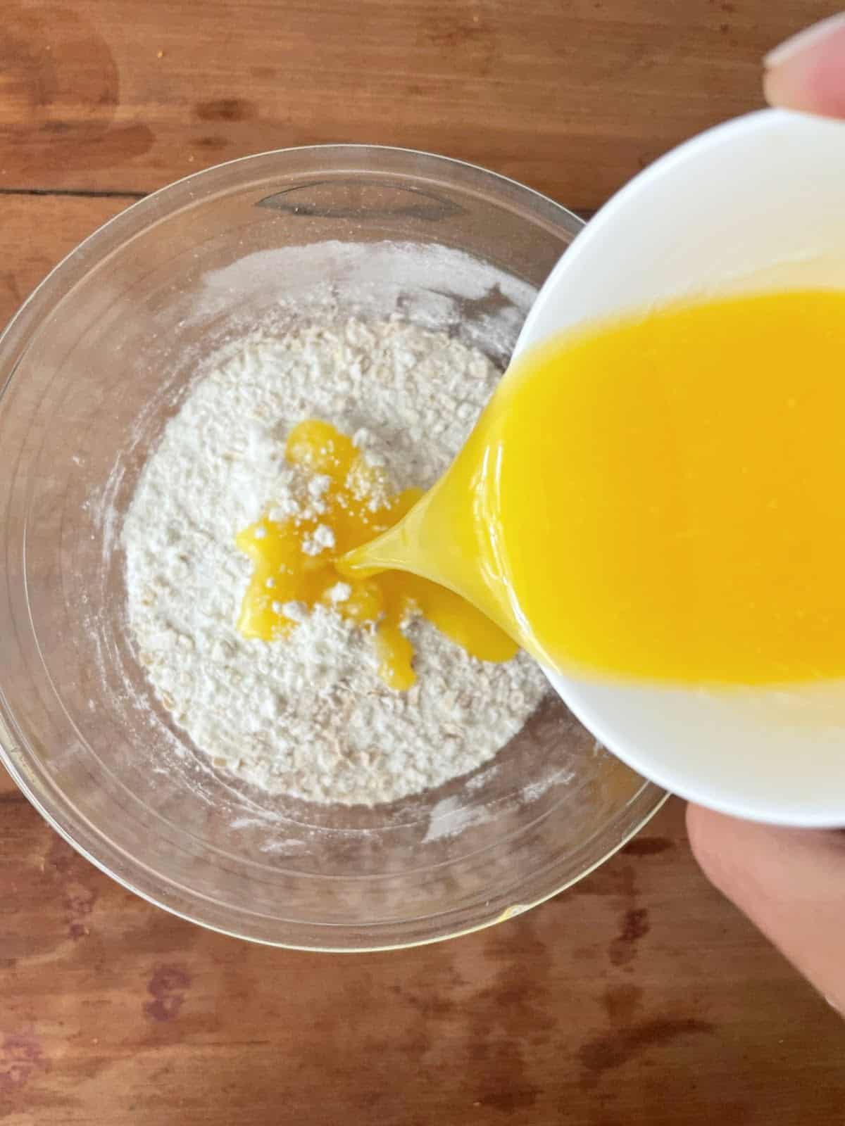 Adding melted butter to dry ingredients in a glass bowl on a wooden table.