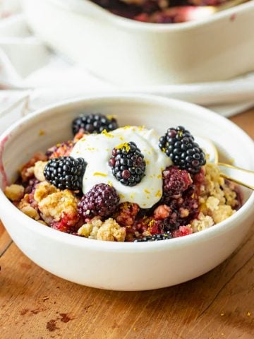 Wooden table with white bowl containing serving of blackberry crumble with whipped cream.