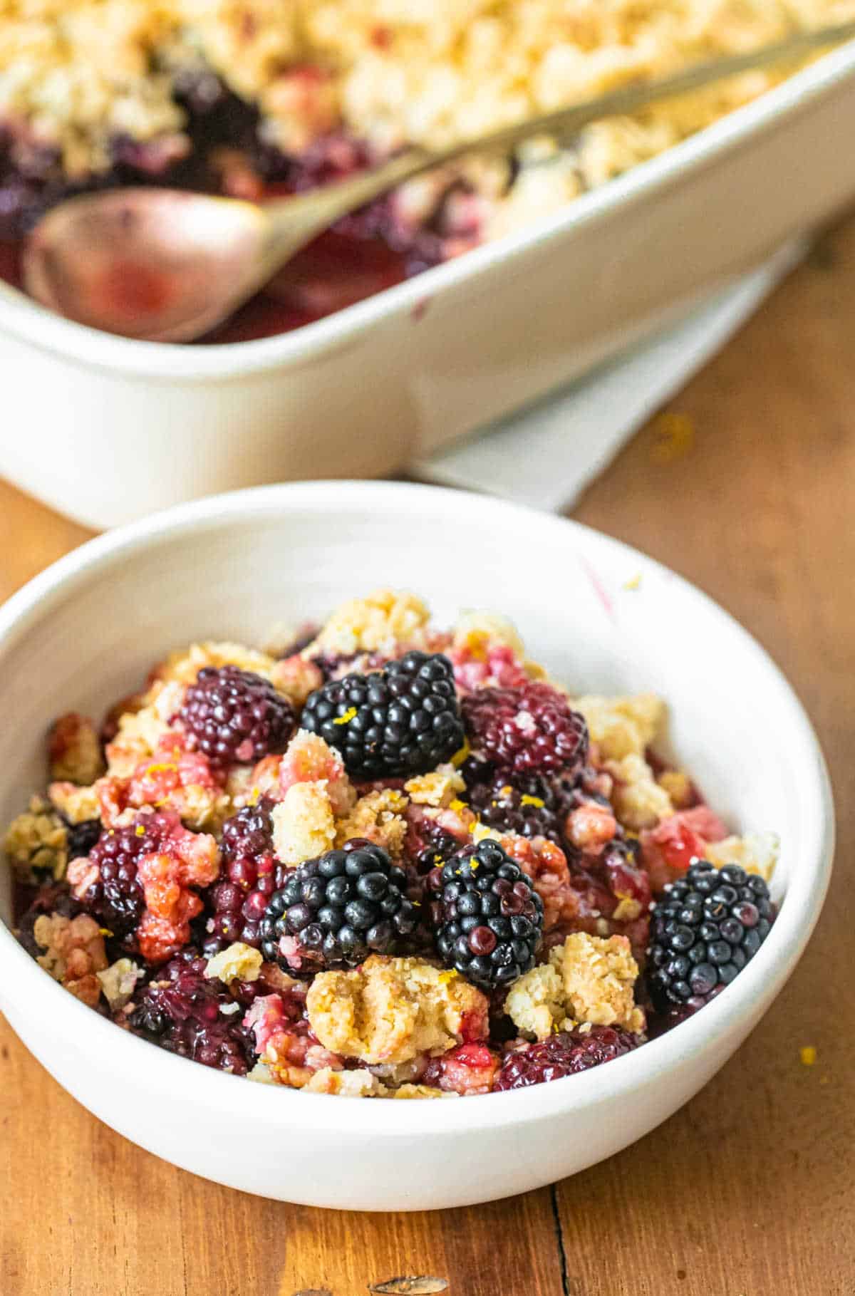 White bowl with blackberry crumble serving on a wooden table. Dish in background.