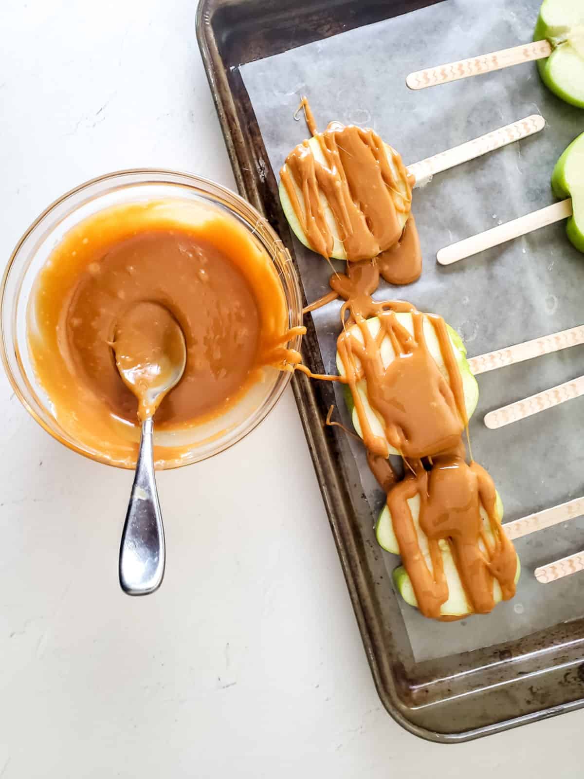 Caramel drizzled apple slices on a baking tray, bowl with melted caramel, white surface.