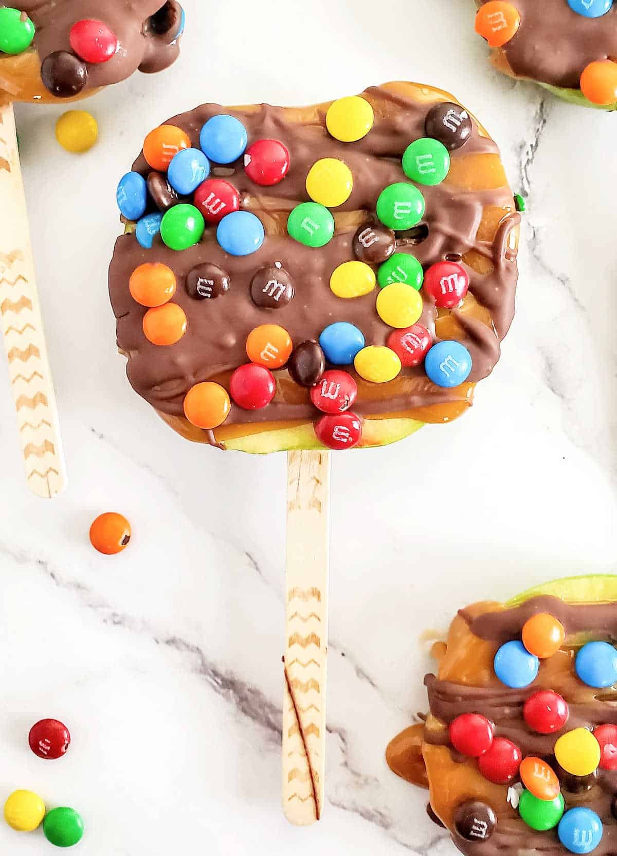 Close up of apple slice on a stick on white marble covered in chocolate and candies.