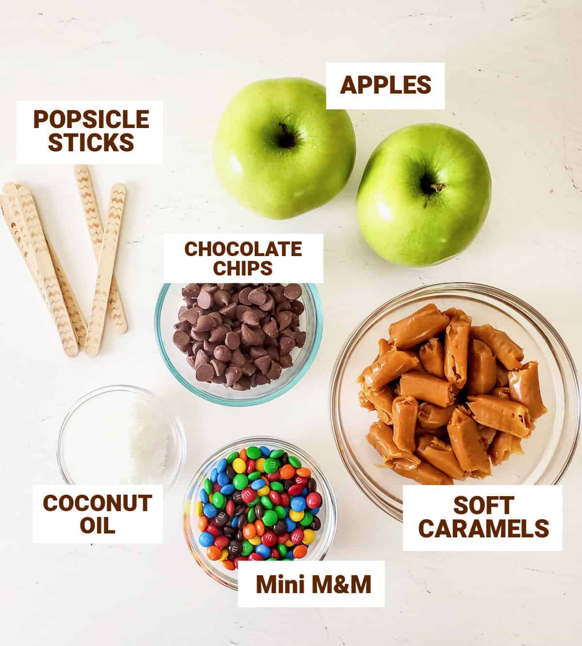 White surface with ingredients for candy apple slices including caramels, chocolate, popsicle sticks, coconut oil.