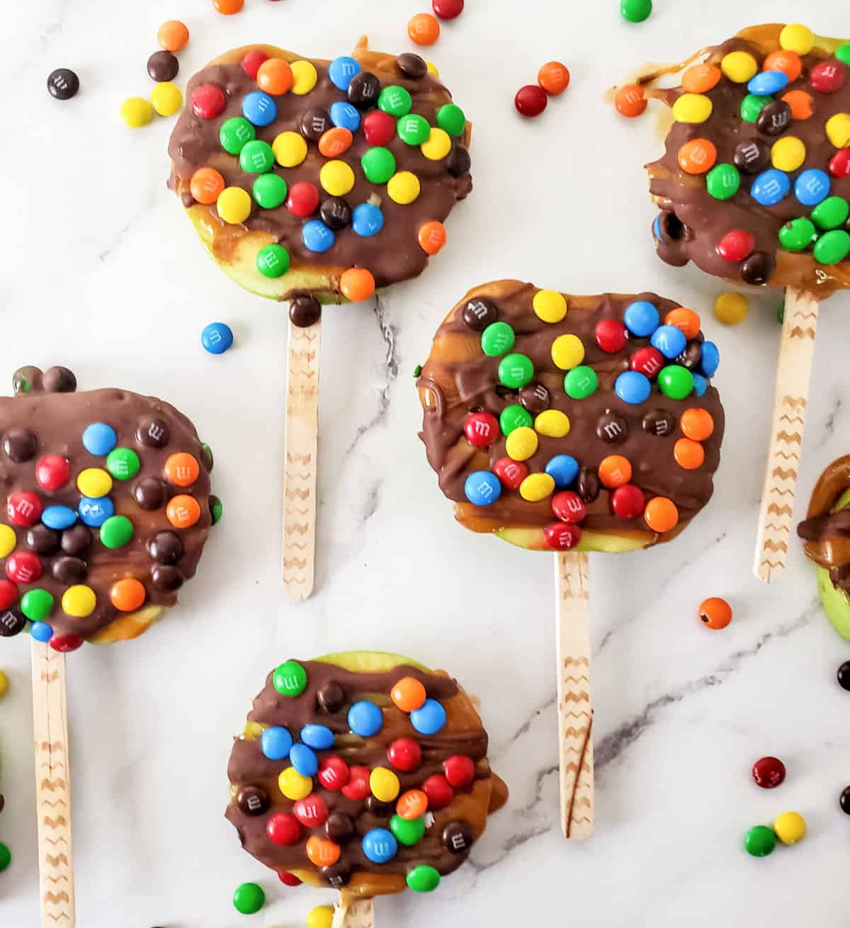 White marble surface with several candy and chocolate covered apple slices on a stick. 