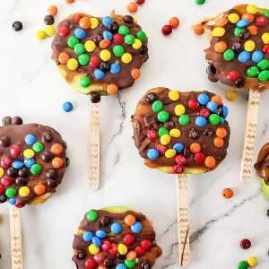 Candy and chocolate covered apple slices on sticks on a white marble surface.