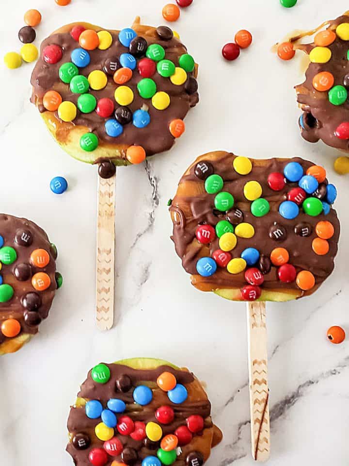 Candy and chocolate covered apple slices on sticks on a white marble surface.