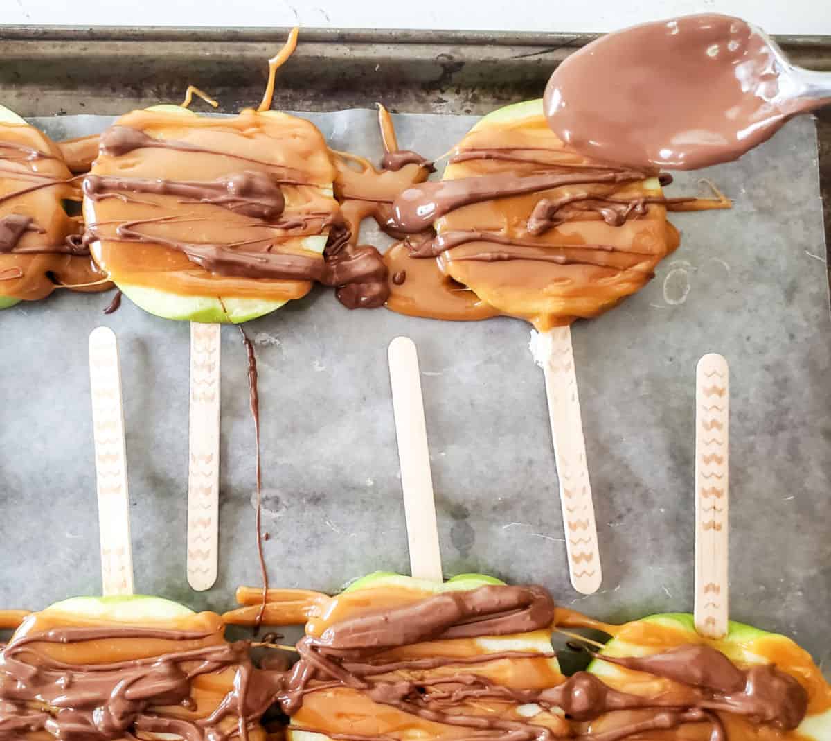 Drizzling chocolate over apple slices on parchment paper on a baking tray.