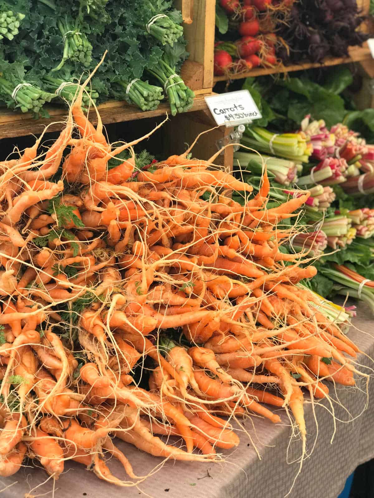 Bunch of carrots on table at farmers market with more vegetables in background.