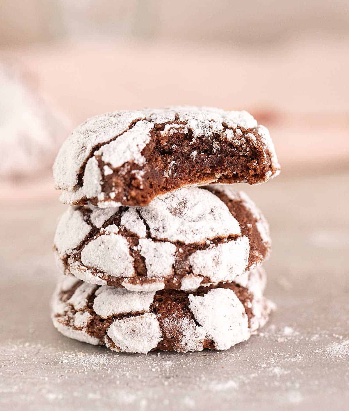Stack of three chocolate crinkle cookie, top one bitter, on grey surface with pink background. 