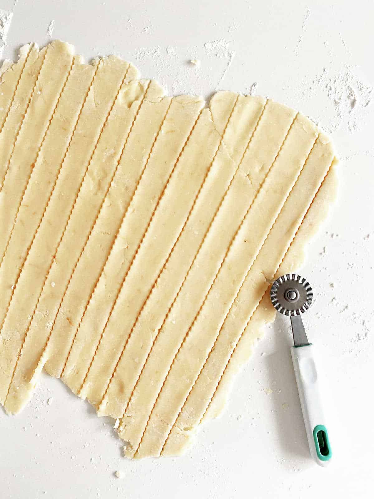 Strips of pie crust on a white surface with pizza cutter on top.