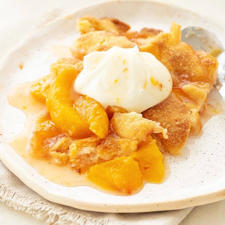 White plate with serving of peach cobbler with whipped cream.