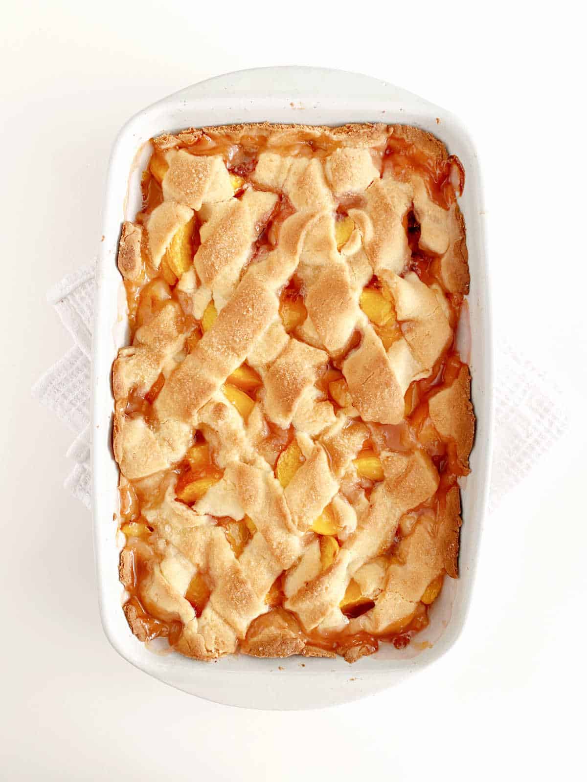 Baked peach cobbler with pie crust lattice in a white ceramic rectangular dish on a white surface.