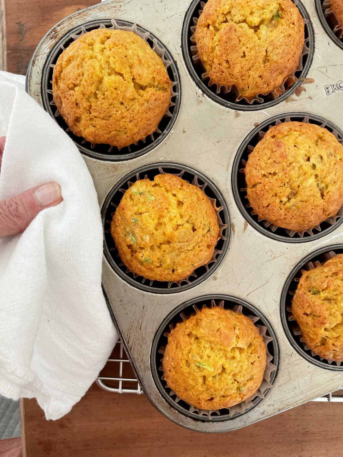 Hand holding baked pumpkin zucchini muffins in a metal pan on a wooden surface with a white kitchen towel.