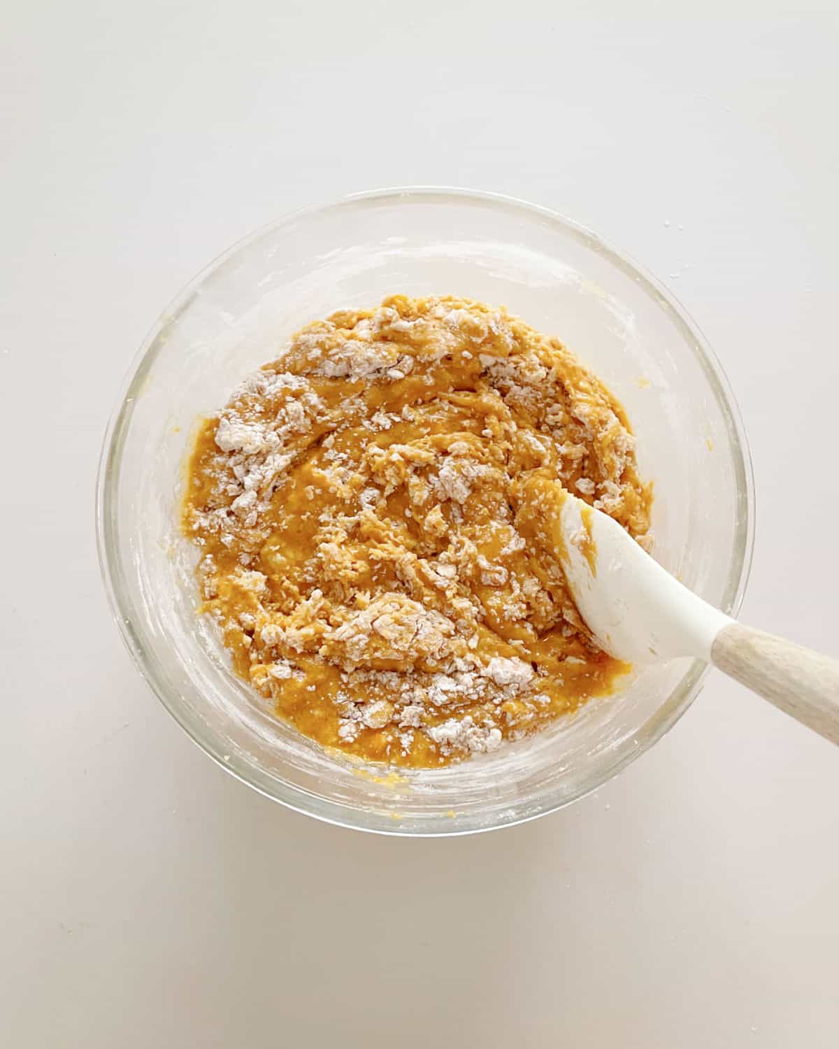 Stirring flour into pumpkin muffin mixture with white spatula in a glass bowl. White surface.