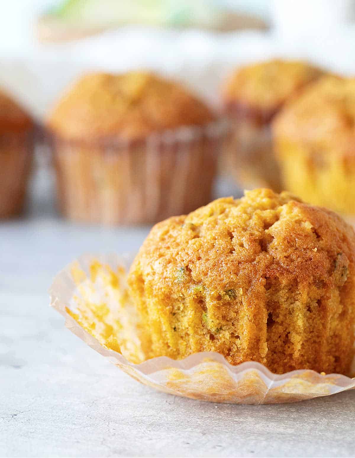 Close up of eaten pumpkin zucchini muffin on a white surface with more muffin in the background.