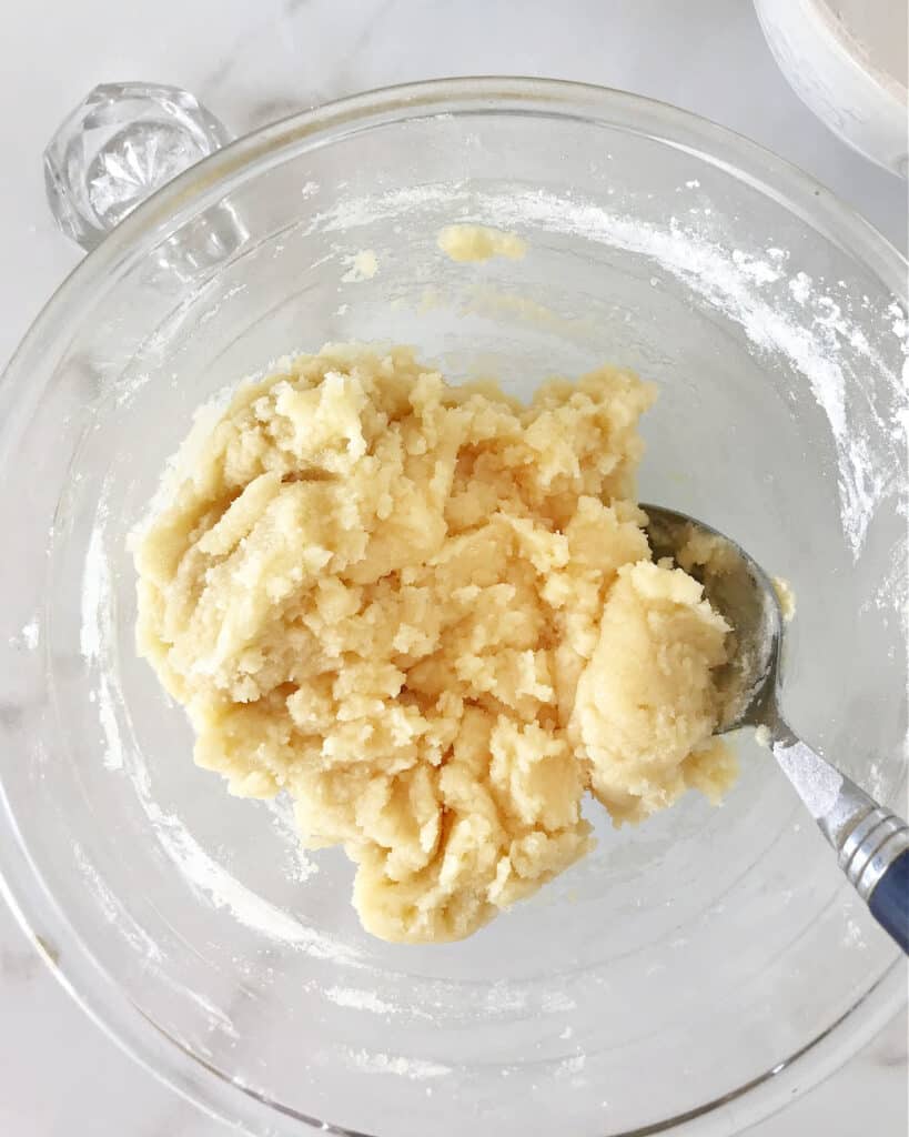 A glass bowl with pie dough mixture and a spoon. White marble surface.