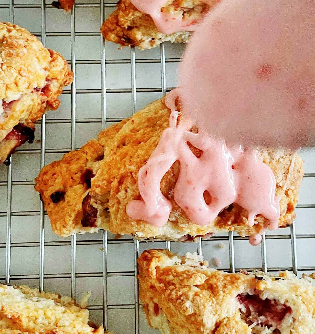 Close up view of strawberry glaze being drizzled on scones on a wire rack.