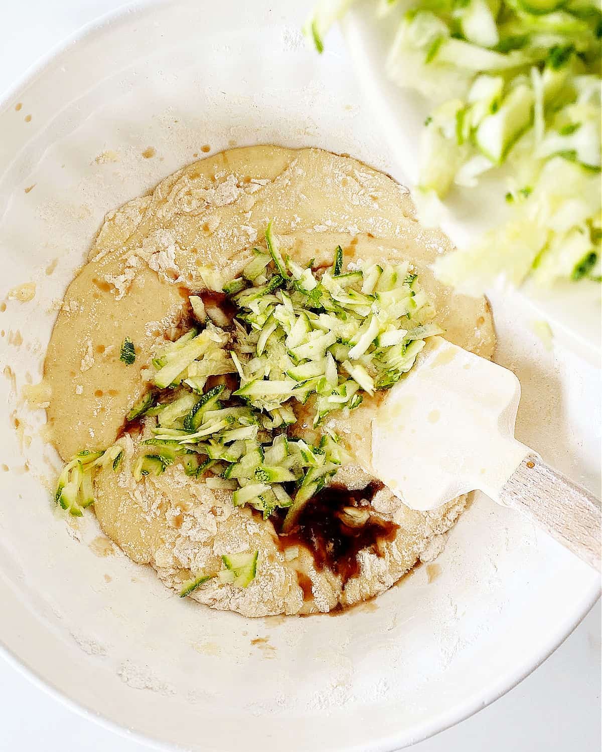 Adding grated zucchini to cake batter in a white bowl.