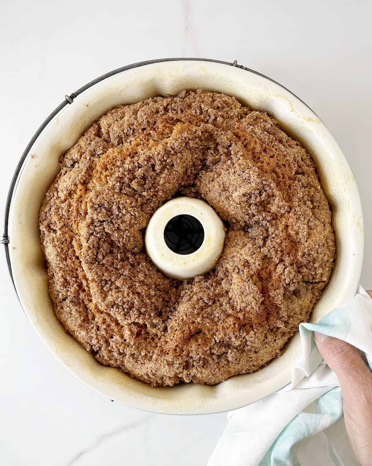 Holding a streusel topped bundt cake in the pan. White marbled surface.