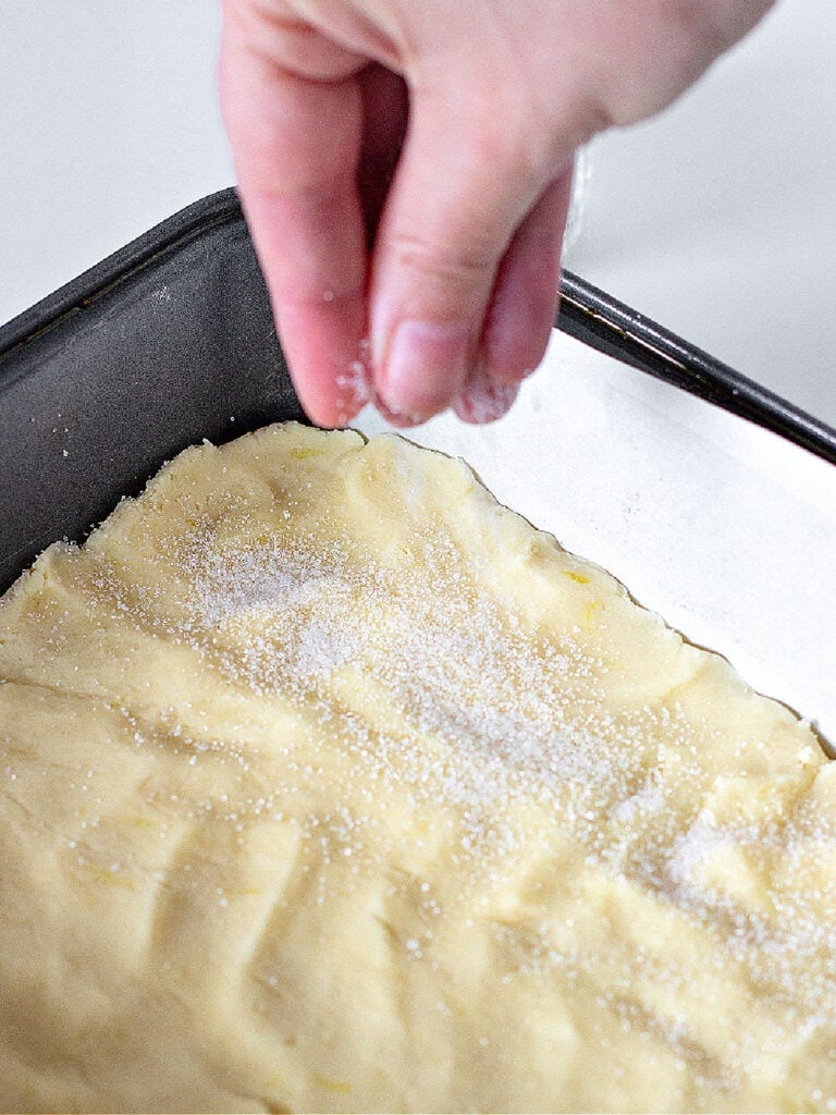 Sprinkling sugar on patted lemon shortbread. Dark metal pan.