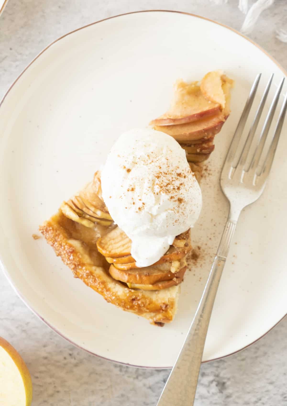A slice of apple tart with cream on a white plate. A silver fork. Grey surface.