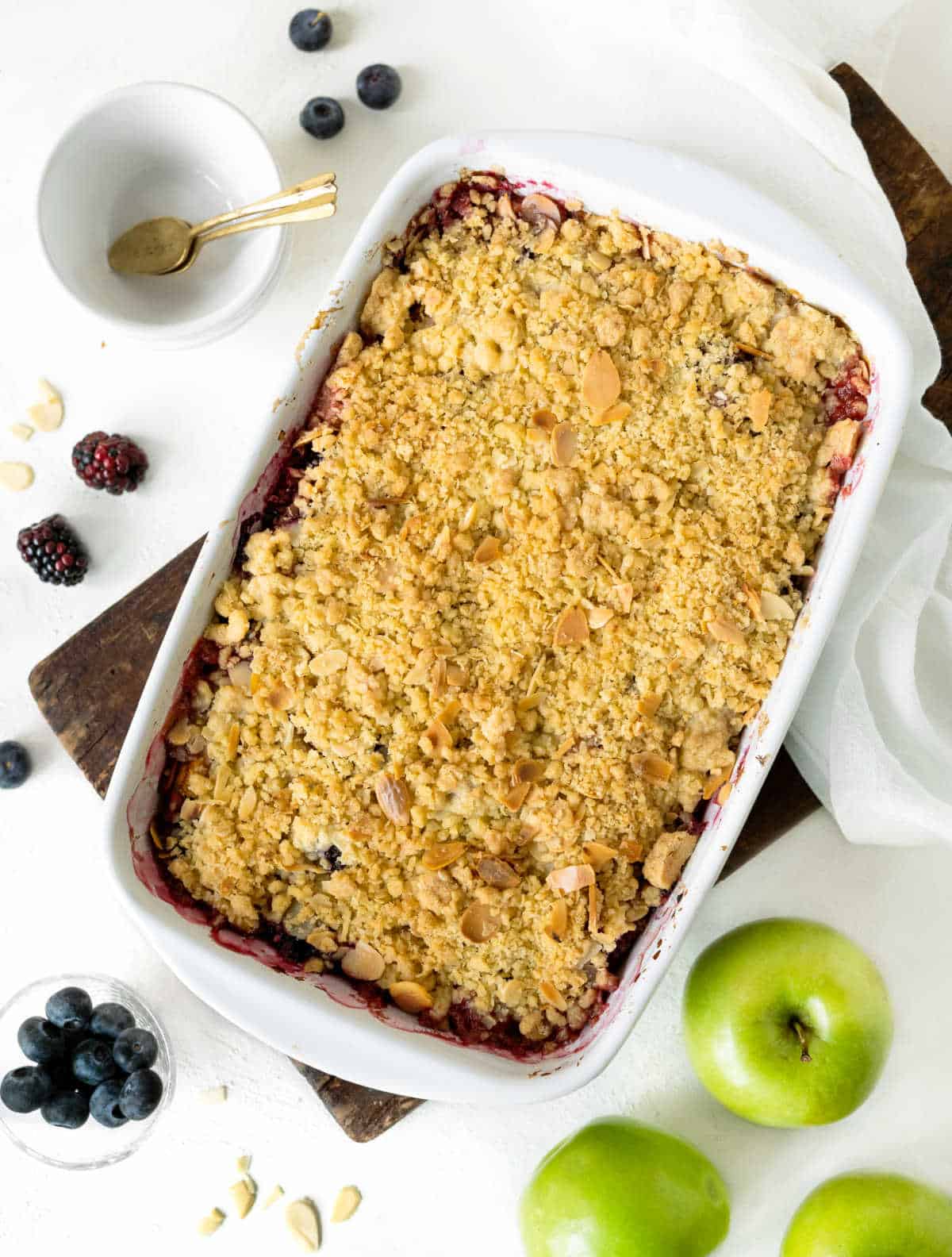 Top view of white baking dish with apple berry crumble on a white surface. Apples, berries, white cloth and bowl around. 