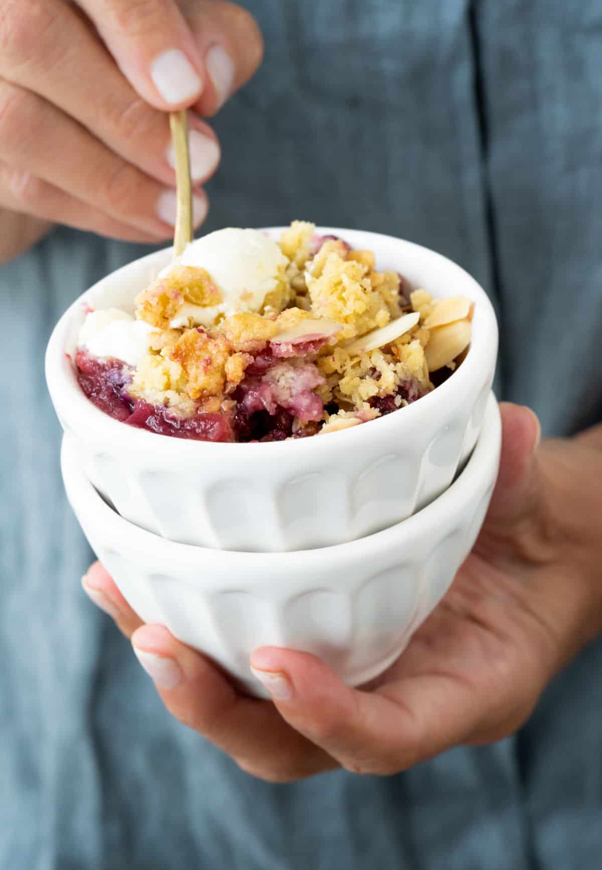 Two white bowls stacked with a serving of apple berry crumble. Hand holding a spoon inside.
