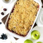 A white bakind dish with apple berry crumble on a white surface. Apples and berries around it. Top view.