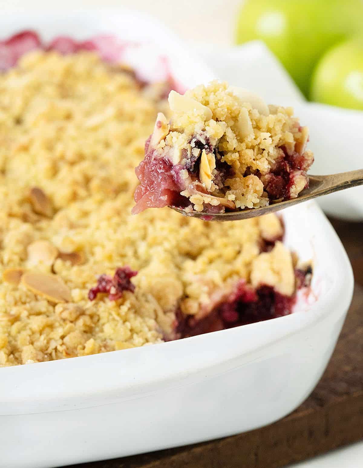 Spoon with apple berry crisp serving over white dish with rest of the dessert on a dark board. Apples in the background.