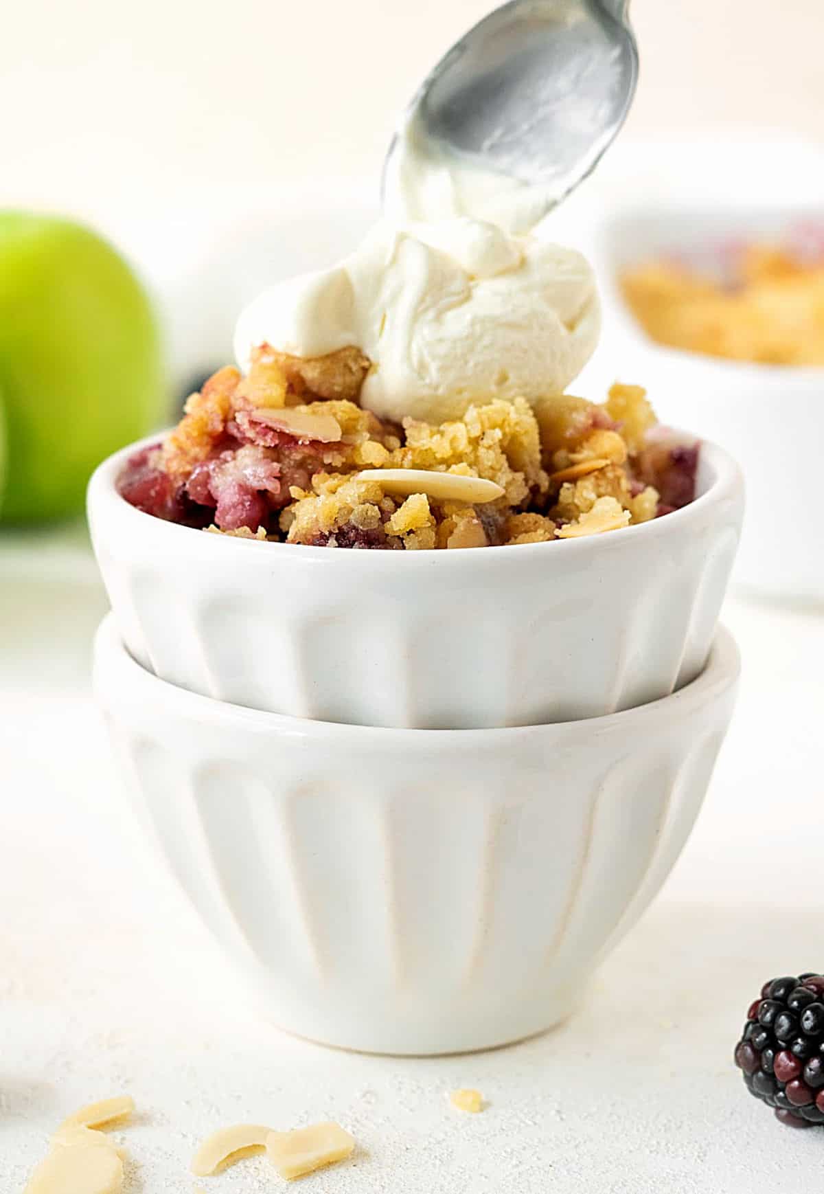 Whipped cream being spooned over stacked white bowls with apple berry crisp. White surface and background.