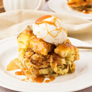 Serving of apple bread pudding with cream and caramel on a white plate.