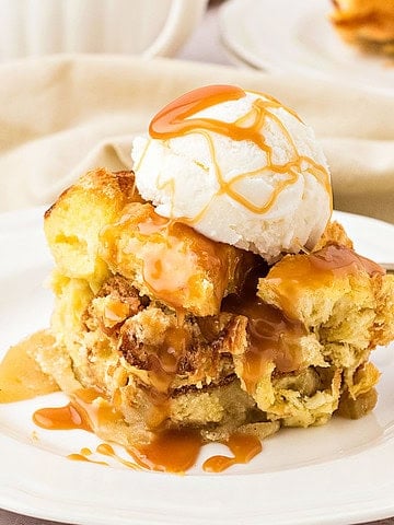 Serving of apple bread pudding with cream and caramel on a white plate.