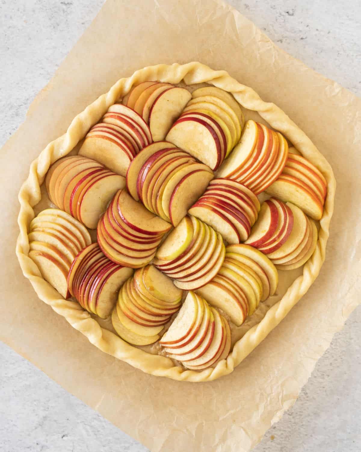 Sliced apples arranged over pie crust to make an apple galette. Beige parchment paper, grey surface.