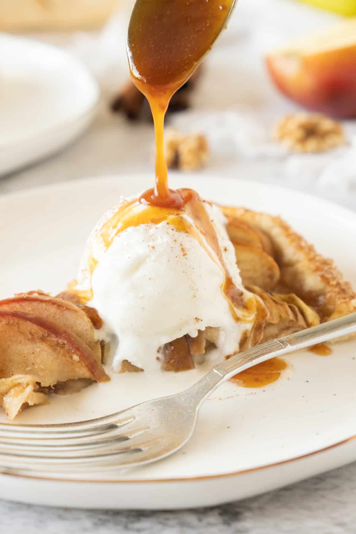 Pouring caramel over slice of apple galette a la mode on a white plate. A silver fork. 