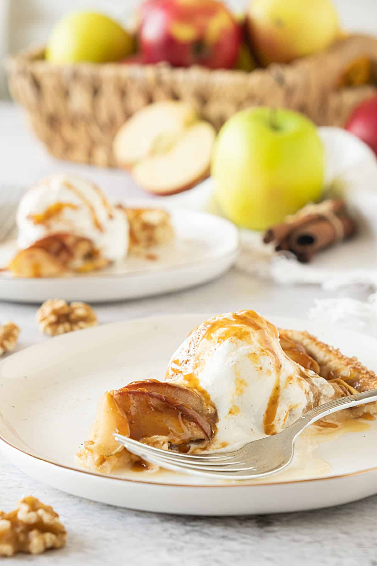 White plates with caramel apple galette slice with ice cream. Basket of apple in the background. Light grey surface. 