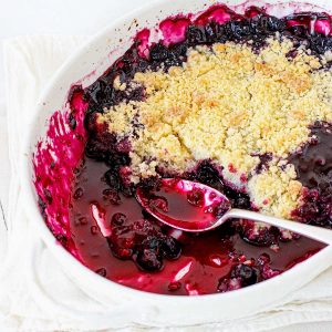 Close up of white oval dish with eaten blueberry crumble with silver spoon. White background.