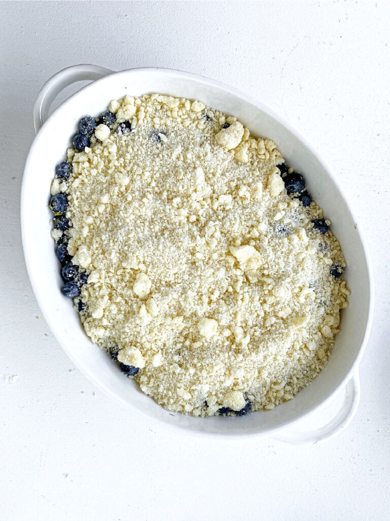 Oval white dish with blueberry crumble before baking. White surface.