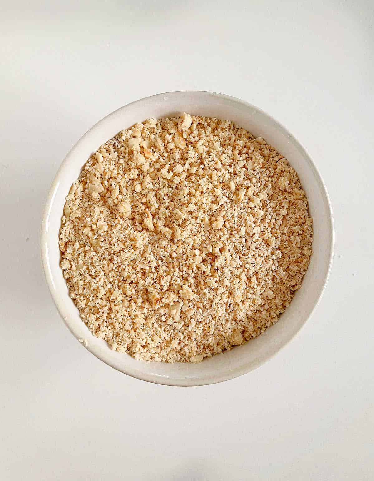 White bowl with ground hazelnuts on a white surface. Top view.