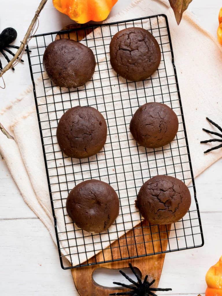 Baked chocolate soft cookies on a black wire rack on a white surface with halloween plastic spiders around.