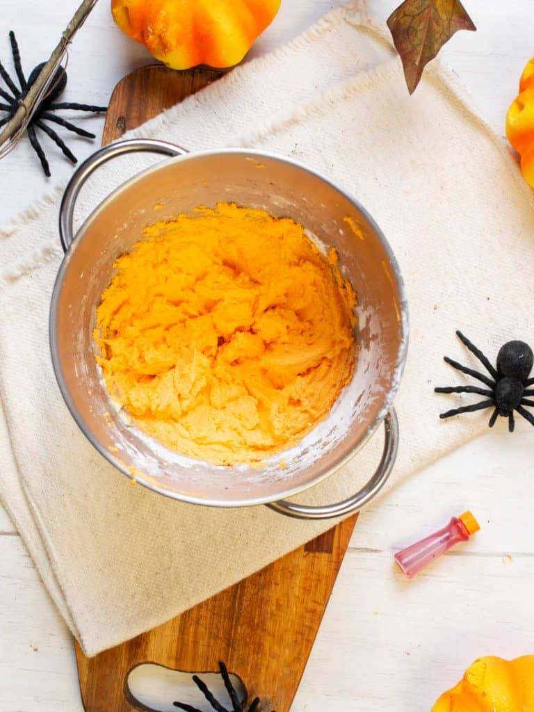 Metal bowl with orange buttercream filling on a white cloth. Black plastic spiders.