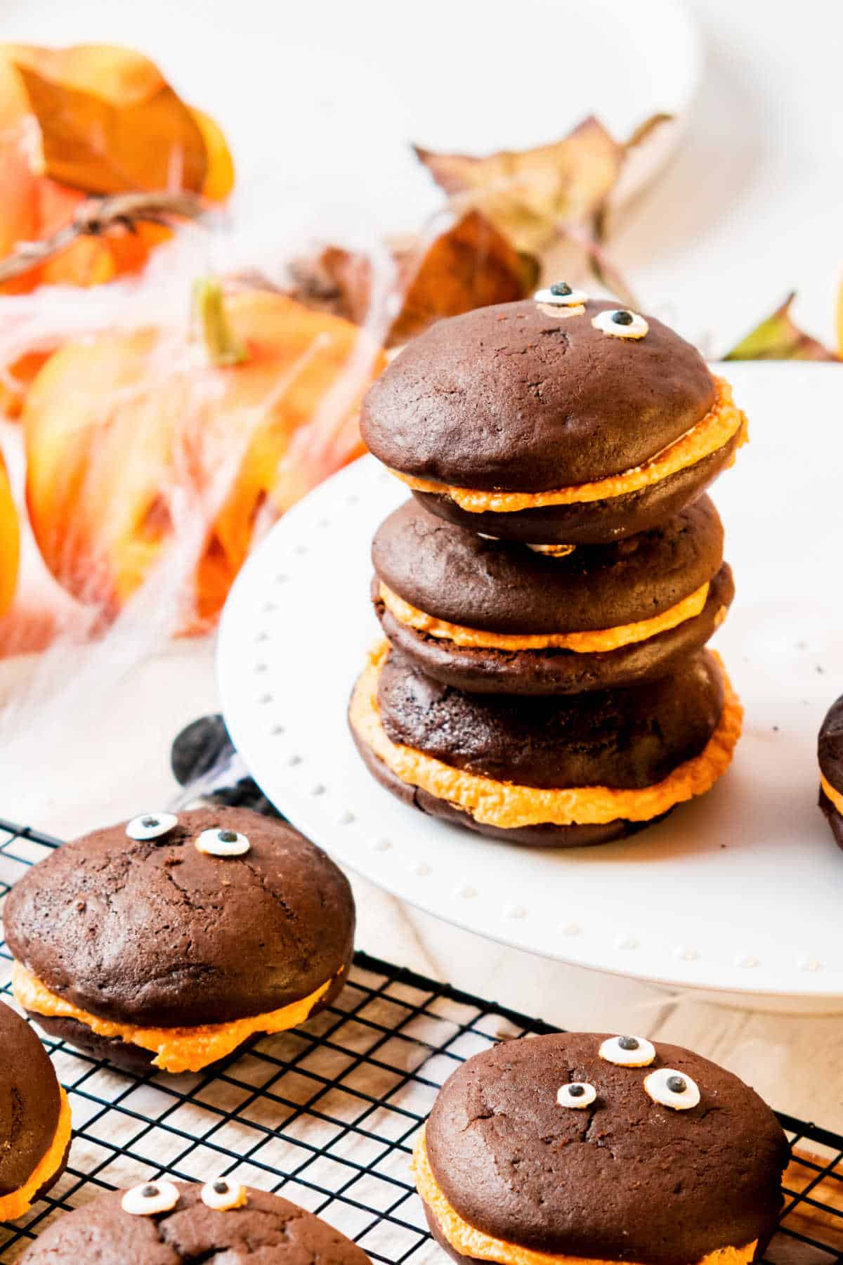 Stack of chocolate whoopies with orange-colored filling on a white plate with halloween props around. 