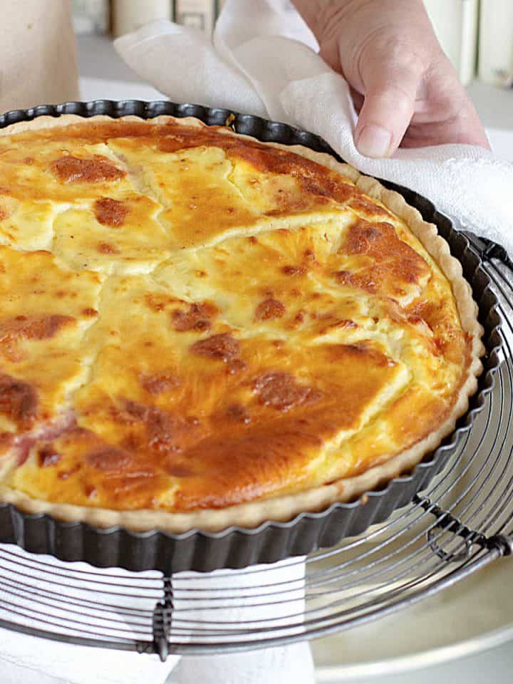 Hands holding whole ham and cheese quiche in a dark metal pan.