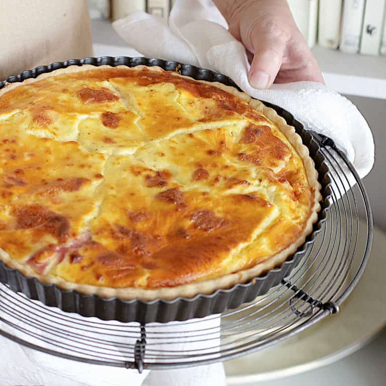 Hands holding whole ham and cheese quiche in a dark metal pan.