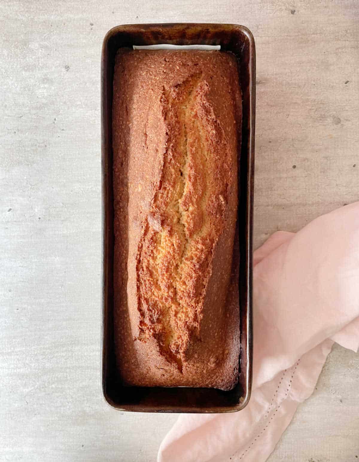 Baked hazelnut loaf cake in a dark metal pan on a grey surface with a pink cloth.