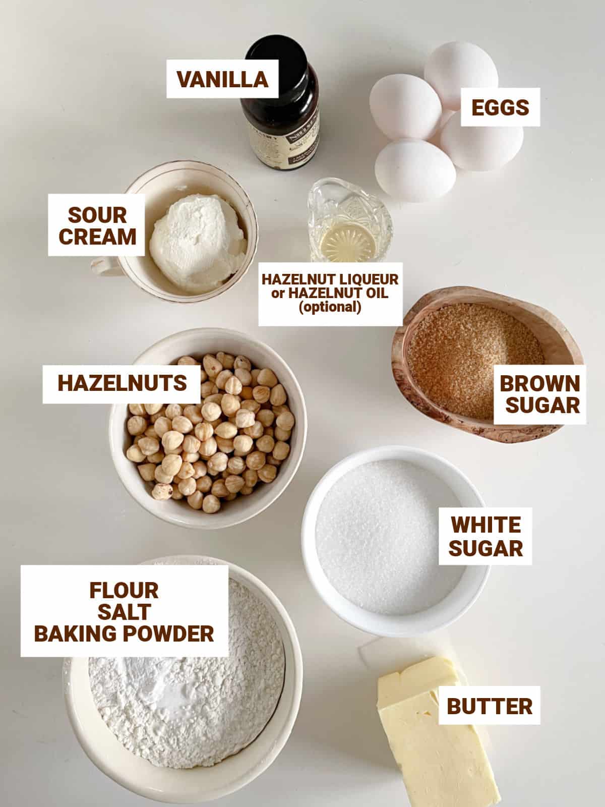 White surface with bowls containing ingredients for hazelnut cake including eggs, sugar, butter, flavorings, flour, sour cream.