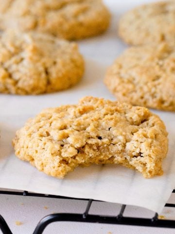 Close up image of bitten oatmeal cookie on white paper on a black wire rack with whole cookies behind.