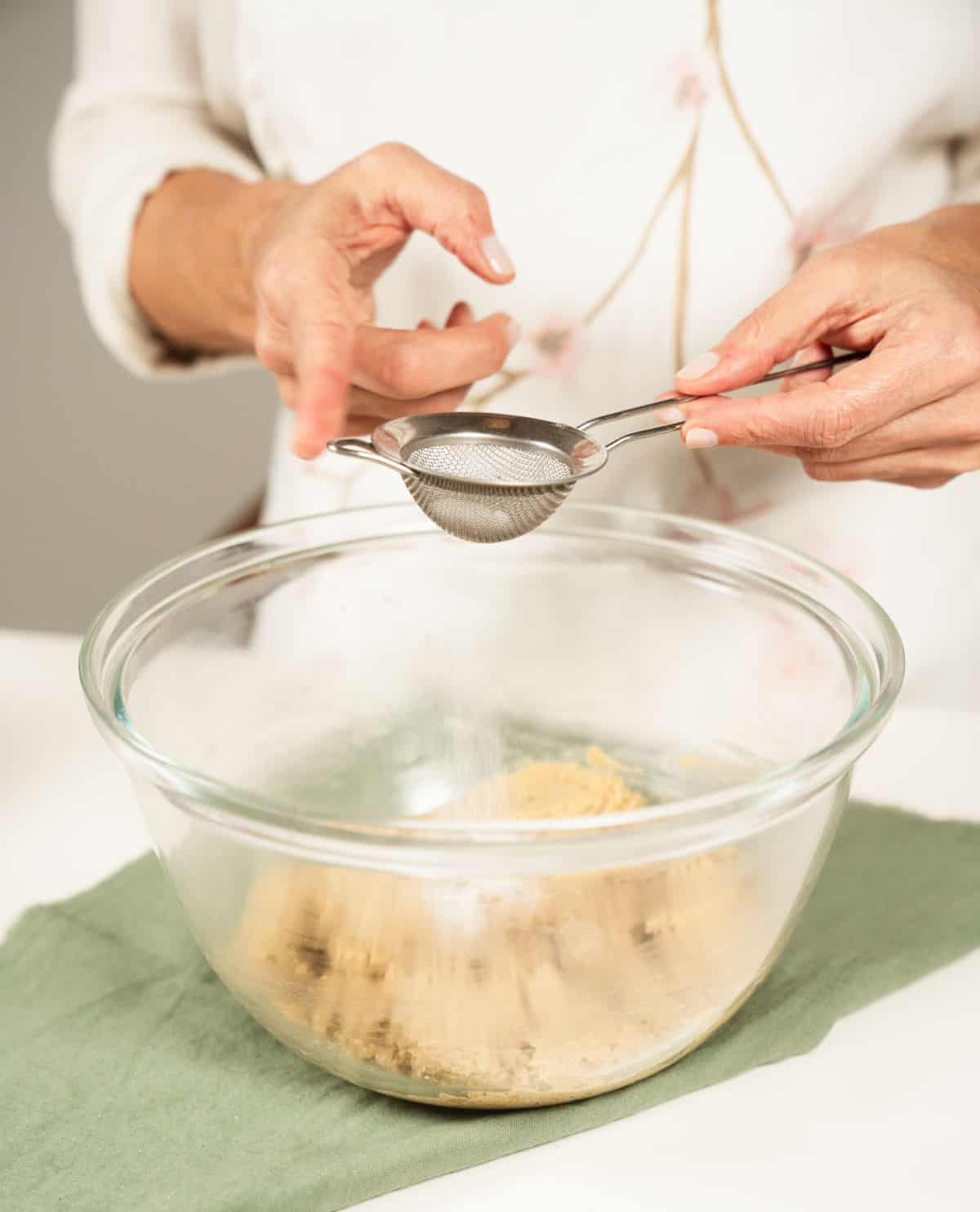 Sifting baking soda over oatmeal cookie dough on a glass bowl with green cloth underneath.